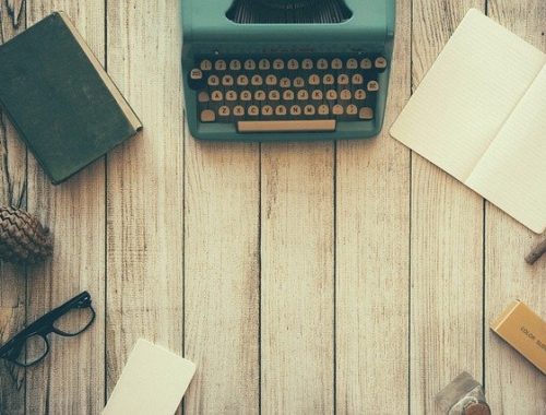 Photo of an old book, typwriter, journal, twig, ink bottle, paper and reading glasses