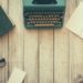 Photo of an old book, typwriter, journal, twig, ink bottle, paper and reading glasses